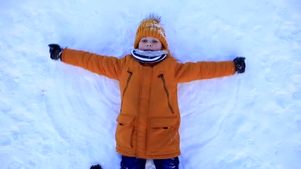 Młody chłopiec w żółtej kurtce leży w śniegu i robi śnieżnego anioła. - Materiał filmowy, wideo