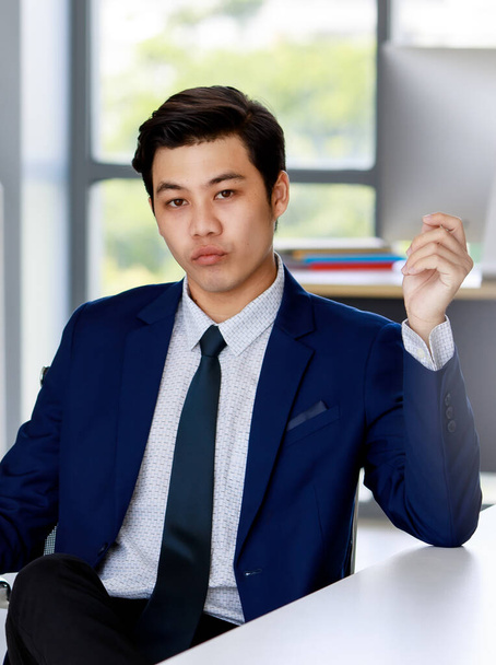 young attractive asian business man wearing navy blue suit with white shirt and necktie sitting down on the executive chair making confident face in office with natural lighting. - Foto, Bild