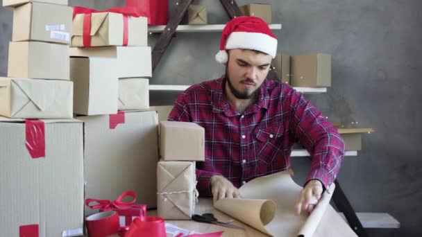 Blanke man in kerstman hoed zit aan de tafel in zijn kantoor en wikkelt een doos in ambachtelijk papier. Kerstverkoopconcept. - Video