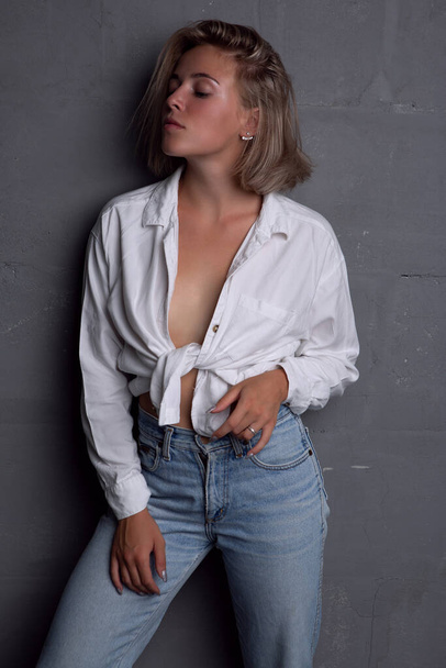 Sexy young blonde woman with a beautiful figure in jeans and an unbuttoned shirt on a gray background - Foto, imagen