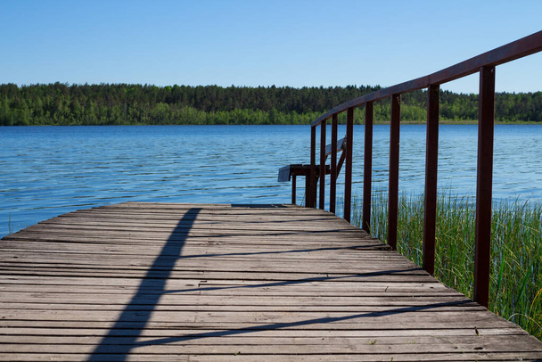 Egy fahíd korláttal és egy pad vezet egy kék tóba, kilátással az erdőre és a kék égre.. - Fotó, kép