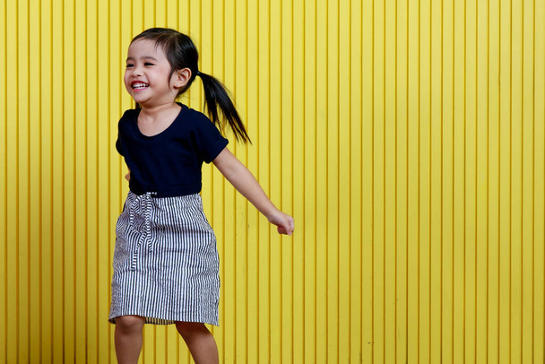 Портретний студійний знімок маленької милої красивої зачіски з косичками дошкільнята щаслива модель дівчини носить чорну сорочку і білу спідницю, що стоїть, посміхаючись позує перед жовтим фоном стіни
. - Фото, зображення