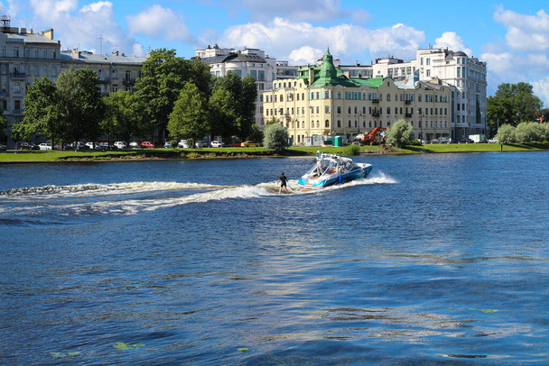  La ragazza è impegnata in wakesurf sul fiume nel parco sotto la guida di un allenatore su una barca sullo sfondo di case e il cielo blu. - Foto, immagini