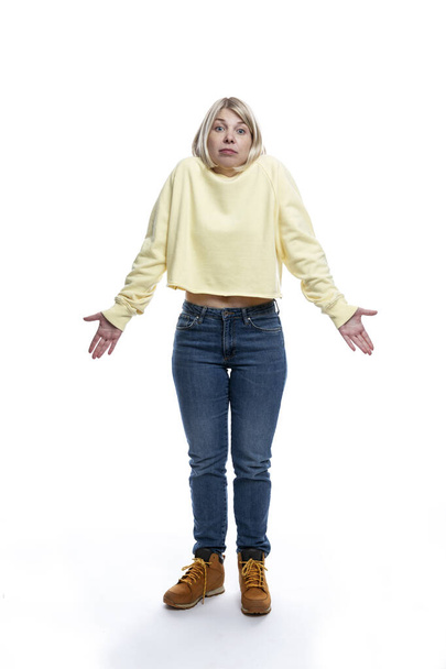 A young woman stands and throws up her hands in surprise. Blonde in a yellow sweater and jeans. Full height. Isolated on white background. Vertical. - Photo, image