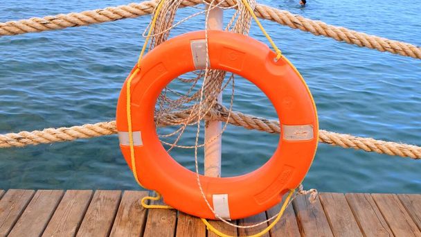 Оранжевый спасательный круг и море на фоне моря и голубого неба. Морские канаты и спасательный жилет висят на столбе. Концепция помощи и безопасности - Фото, изображение