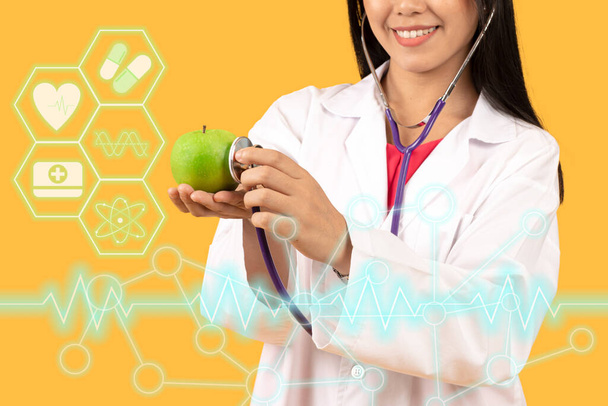 Pankart ve seçici odak noktası, beyaz önlük giyen, yeşil elmayı kontrol etmek için steteskop kullanan, turuncu arka planda tıbbi servis simgeleri olan, gülen, sağlıklı kadın doktorların elleri. - Fotoğraf, Görsel