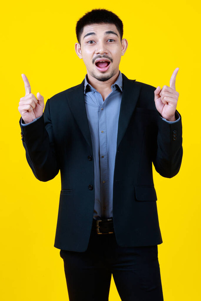 Asian business beard handsome adult man wearing formal suit, raising hands, presenting and pointing up to above copy blank space, making gesture of surprise, standing on isolated yellow background - Foto, afbeelding