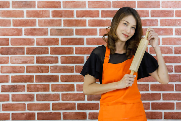 Όμορφη νεαρή Ασιάτισσα γυναίκα σε πορτοκαλί ποδιά χαμογελώντας στέκεται κοντά στον τοίχο από τούβλα και αστεία στάση από αστειεύεστε δείχνει ξύλινο πλάστη στο σπίτι κουζίνα - Φωτογραφία, εικόνα