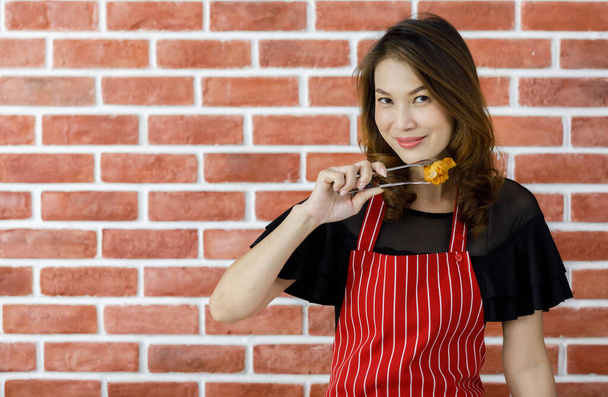 Atractiva linda mujer asiática joven en delantal de rayas rojas sonriendo posar con diversión por el ala de pollo frito sabroso pellizco y mantenerlo cerca de la cara de broma cerca de la pared de ladrillo estampado en la cocina casera - Foto, imagen