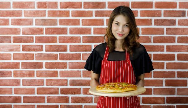 Preciosa mujer asiática joven como chef en delantal de rayas rojas de pie cerca de la pared de ladrillo y sonriendo levantando un plato de madera de sabrosa pizza cocida para persuadir a comer deliciosa comida italiana tradicional - Foto, imagen