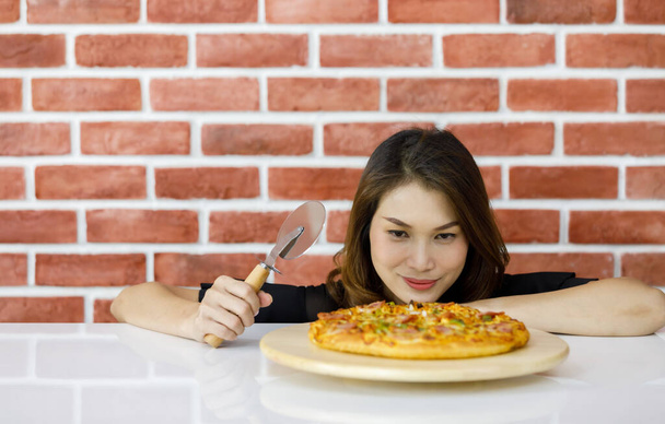 Kaunis nuori aasialainen nainen alempana lähellä tiiliseinää ja hymyillen katsoen hyvin lähelle houkuttelevaa maukasta ja terveellistä tuoretta pizzaa, kun hänen rakastamansa italialainen ruoka pyöreällä puisella lautasella valkoisella pöydällä - Valokuva, kuva
