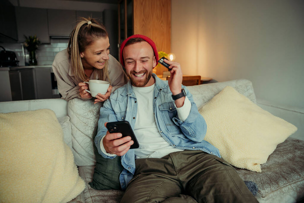 Kaukasisches Paar sitzt auf Couch und trinkt Kaffee und bezahlt online mit Mobiltelefon - Foto, Bild