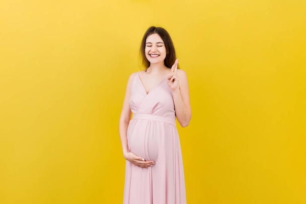 Cheerful young pregnant woman holding fingers crossed, waiting pregnancy isolated on colored background in studio. lifestyle pregnancy concept. - Photo, image