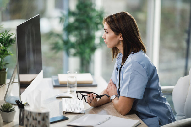 Una atractiva enfermera joven usando una computadora mientras está sentada en su escritorio en el hospital. - Foto, Imagen