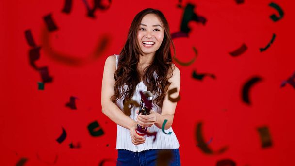 Funny young and cure Asian girl holding and blowing out party poppers with funny and happy. Studio shot on red background. - Photo, Image