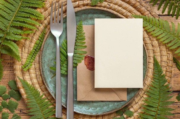 Blank paper card and sealed envelope on table setting decorated with fern leaves on wooden table top view. Tropical mock-up scene with invitation card flat lay - Foto, immagini