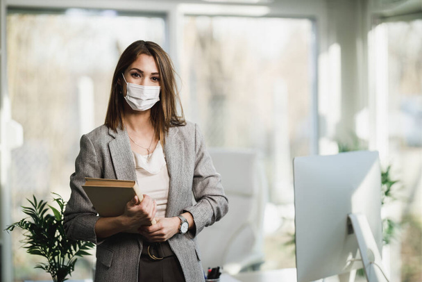 A successful young business woman with surgical mask standing in her office. Looking at camera. - Fotoğraf, Görsel