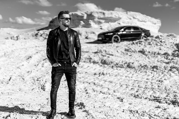 Brutal man wearing suit is standing at the desert near his car. Man in front of the luxury auto. Horizontal photo - Фото, изображение