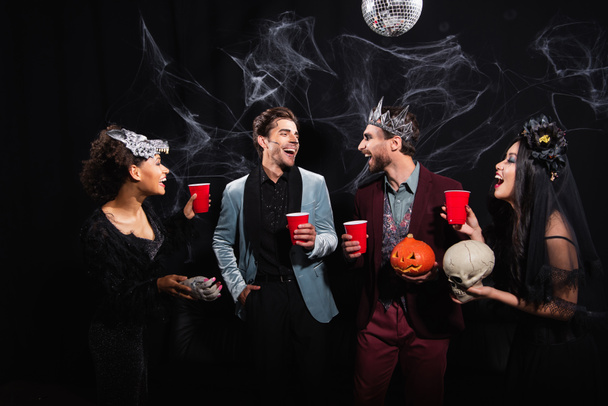 happy multiethnic friends with plastic cups talking during halloween party on black  - Photo, Image