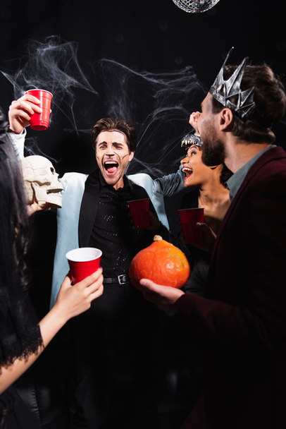hombre riendo sosteniendo taza de plástico cerca de amigos multiétnicos en trajes de Halloween en negro - Foto, Imagen