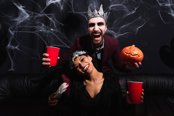laughing man in vampire halloween costume with plastic cup and carved pumpkin near african american woman in wolf mask on black   - Photo, Image