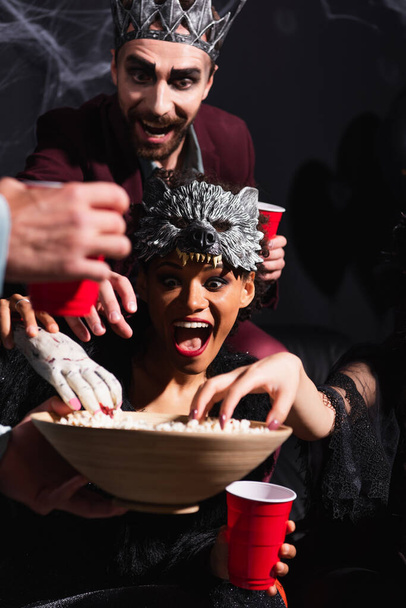 vzrušený africký americký žena ve vlk maska přičemž popcorn s děsivý hračka ruka v blízkosti přátel na černé - Fotografie, Obrázek