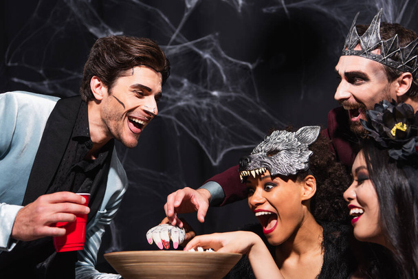 удивленные многонациональные друзья едят попкорн во время Хэллоуина партии на черном - Фото, изображение