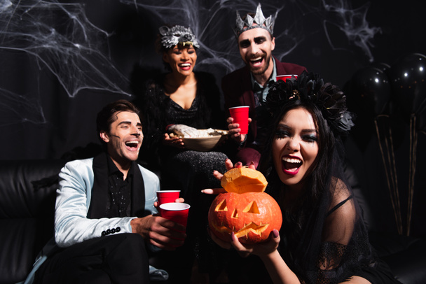 aufgeregte Asiatin zeigt geschnitzten Kürbis neben lachenden multikulturellen Freunden in Halloween-Kostümen auf schwarz - Foto, Bild