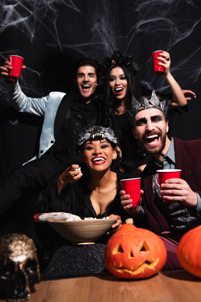 cheerful multiethnic friends in halloween costumes looking at camera while eating popcorn and drinking beer on black - Fotó, kép