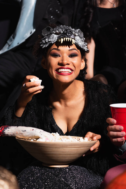 excited african american woman in wolf mask eating popcorn during halloween party on black - Photo, Image