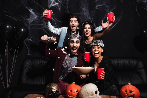 amis multiculturels excités en costumes d'Halloween près de pop-corn, citrouilles sculptées et crânes sur noir - Photo, image