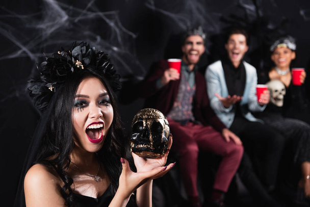 Azjatka w stroju wampira halloween, ze straszną czaszką, warczy przed kamerą w pobliżu rozmazanych przyjaciół na czarno - Zdjęcie, obraz