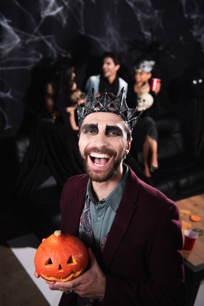 cheerful man in vampire halloween costume laughing at camera while holding carved pumpkin on black - Valokuva, kuva