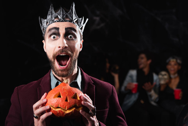 opgewonden man in vampier koning kroon houden griezelige halloween pompoen terwijl kijken naar camera op zwart - Foto, afbeelding