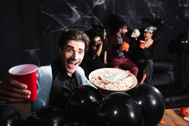 feliz hombre en halloween maquillaje celebración de cerveza y palomitas de maíz cerca de globos negros y amigos interracial sobre fondo borroso y negro - Foto, imagen