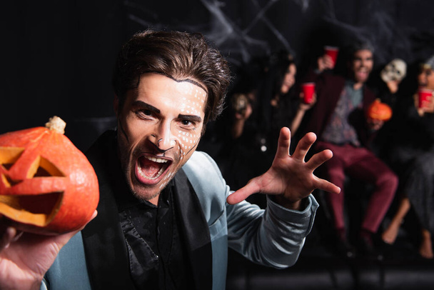 man in halloween makeup growling and showing scary gesture while holding carved pumpkin on black - Foto, Imagem