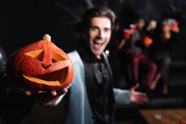 расплывчатый мужчина, держащий жуткую резную тыкву во время вечеринки на Хэллоуин на черном - Фото, изображение