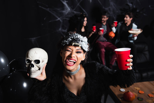 mujer afroamericana en máscara de halloween lobo sobresaliendo lengua mientras sostiene espeluznante cráneo y taza de plástico en negro  - Foto, imagen