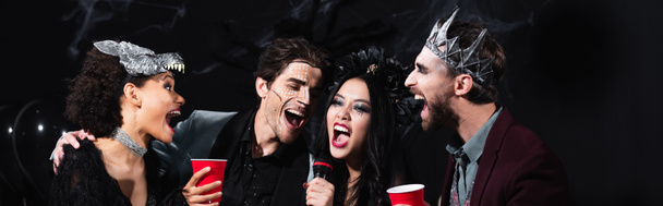 podekscytowani wielonarodowi przyjaciele w kostiumach halloween i makijażu śpiewający karaoke na czarnym, sztandar - Zdjęcie, obraz