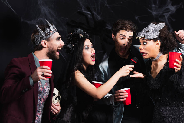amis multiethniques joyeux avec des tasses en plastique chantant karaoké pendant la fête d'Halloween sur noir  - Photo, image