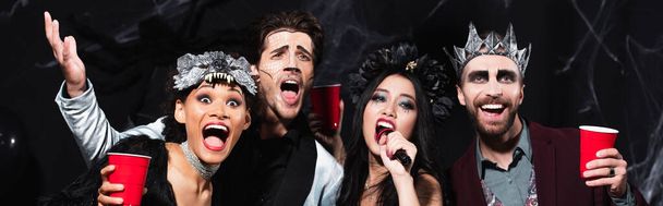 man in halloween makeup gesturing while singing karaoke with multiethnic friends on black, banner - Фото, изображение