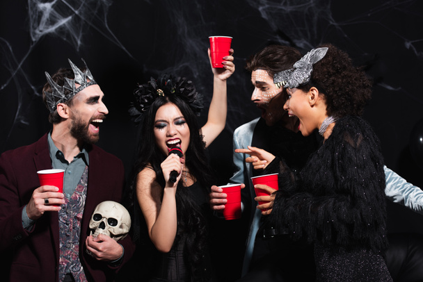 femme asiatique avec tasse en plastique chantant karaoké pendant la fête d'Halloween avec des amis multiethniques sur noir - Photo, image
