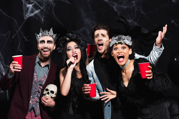 wesoły wielokulturowy przyjaciele w halloween makijaż i kostiumy śpiew karaoke na czarny - Zdjęcie, obraz
