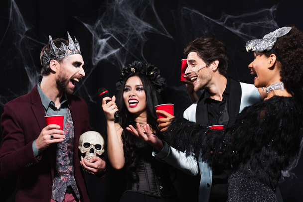 happy asian woman in vampire halloween costume singing karaoke near multiethnic friends on black  - Фото, изображение