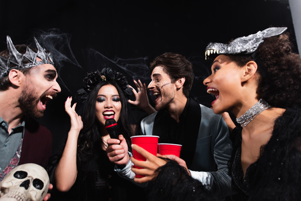 мужчина в макияже на Хэллоуин держит микрофон рядом с азиаткой поющей караоке с друзьями по черному   - Фото, изображение