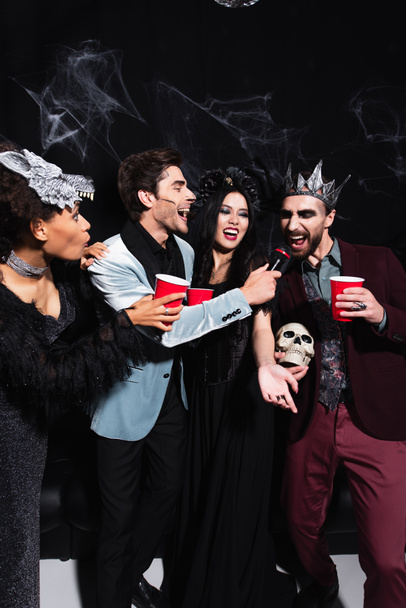 man with skull and plastic cup singing karaoke near multiethnic friends in halloween costumes on black  - Fotografie, Obrázek