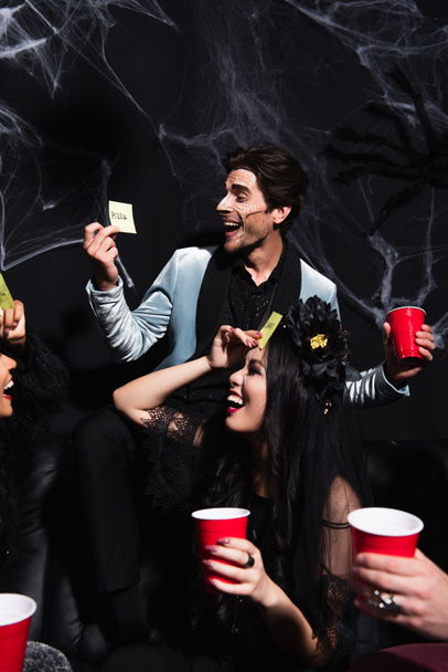 hombre alegre mirando nota pegajosa mientras juega adivina quién juego con amigos multiétnicos en la fiesta de Halloween en negro - Foto, Imagen
