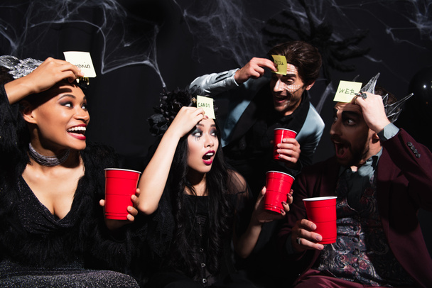 изумленная азиатка рядом с весёлыми многонациональными друзьями, играющими в угадай, кто играет на хэллоуинской вечеринке на черном - Фото, изображение
