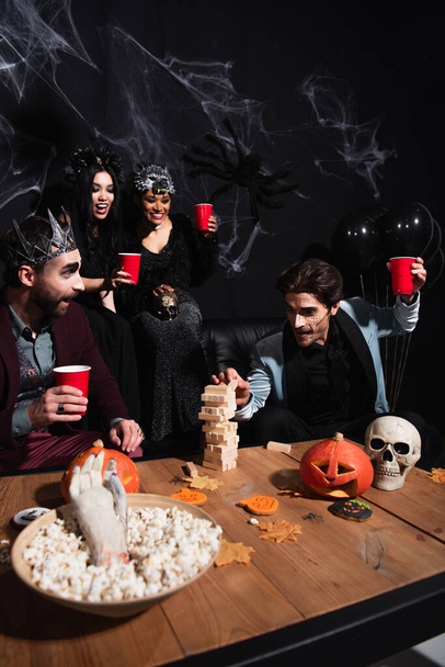 hombre en Halloween maquillaje jugando bloques de madera juego con amigos multiétnicos cerca de calabazas talladas y palomitas de maíz en negro   - Foto, Imagen