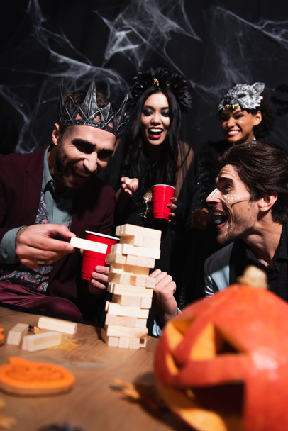 alegre hombre en vampiro rey corona jugando bloques de madera juego durante la fiesta de halloween con amigos multiétnicos en negro   - Foto, Imagen